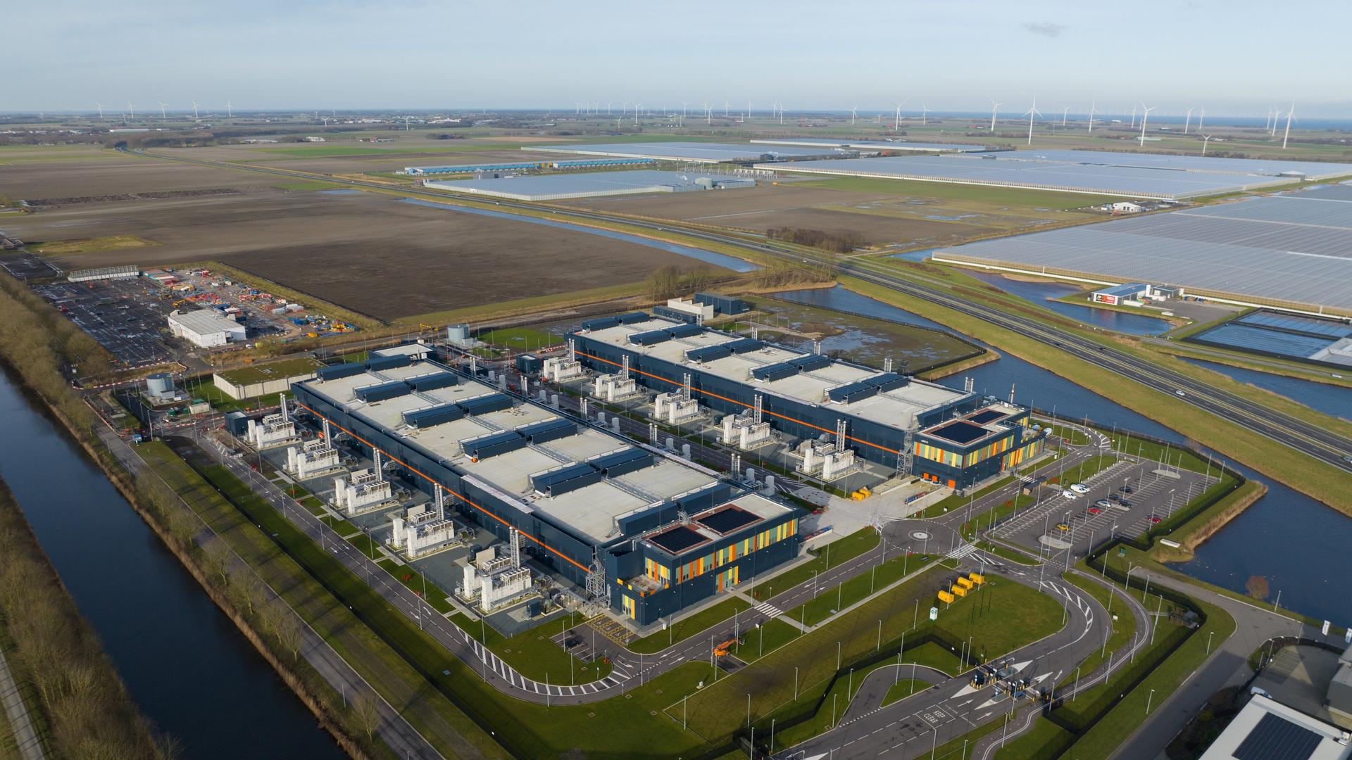 Large data center campus showing multiple buildings and infrastructure, surrounded by canals and agricultural fields, with wind turbines visible in the distance